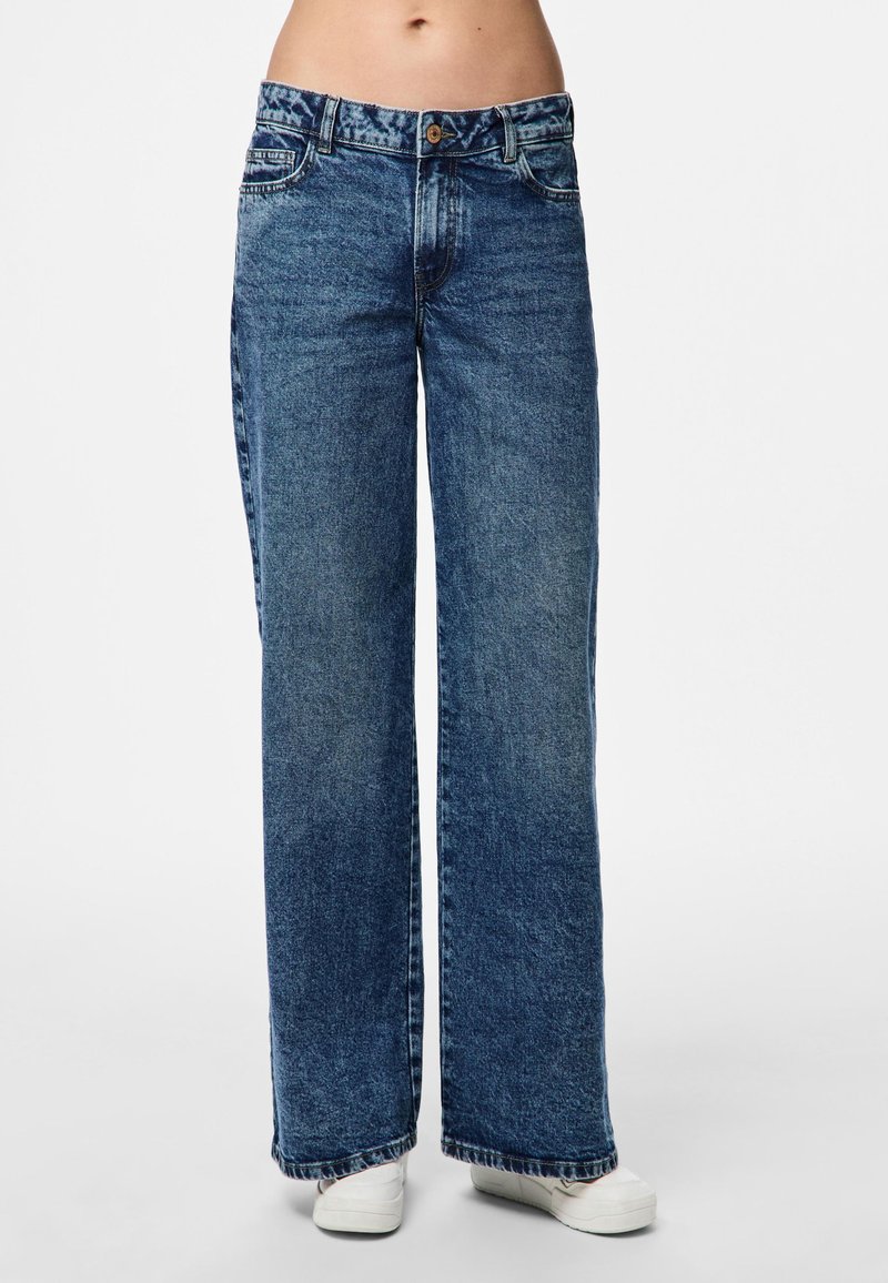 Plati kojinės džinsai vidutinio mėlynumo, turintys mygtuko uždarymą, priekinės kišenės ir nusidėvėjusią tekstūrą. Papuošti prailgintu hemu.