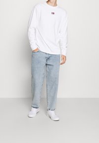 Mann trägt ein weißes Langarmshirt mit Logo, hellblaue, lockere Jeans, weiße Sneaker und steht mit einer Hand in der Tasche vor einem hellen Hintergrund.