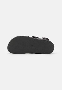 Sort sandal sål med tekstureret gummimateriale. Kontureret form med minimale trækkraftmønstre og lille hardwaredetalje nær stroppen.