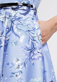 Robe florale bleu clair avec des motifs détaillés en blanc et bleu plus foncé. Elle présente une ceinture noire et une poche latérale pour plus de fonctionnalité.