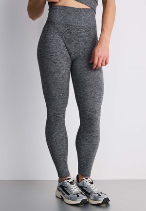 Kobieta ubrana w szare teksturowane legginsy z wysokim stanem oraz srebrno-czarne buty do biegania, stoi z jedną ręką opartą blisko biodra.