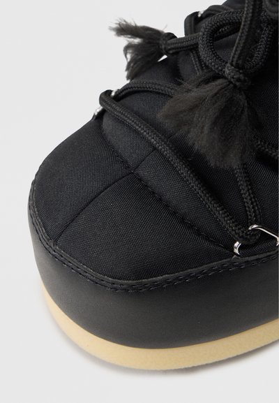 Adidași negri cu un material texturat pe partea superioară, șireturi distinctive cu ciucuri, vârf rotund și o talpă din cauciuc cu margine de culoare deschisă.
