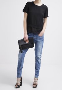 Svart kortärmad topp med genomskinligt underlager; ljusblå slitna jeans; svart väska med fransar; tan färgade remsandaler med klack.