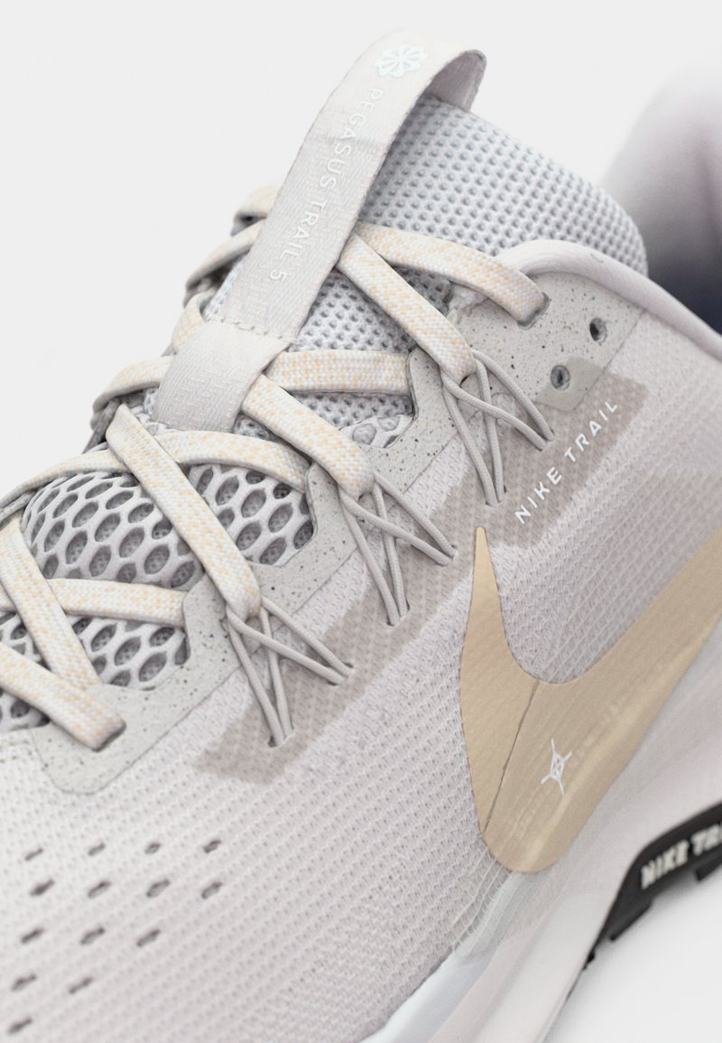 Primer plano del zapato de correr Nike Trail Pegasus Trail 3 en gris claro que muestra la parte superior de malla, los cordones y el logo Nike swoosh beige.