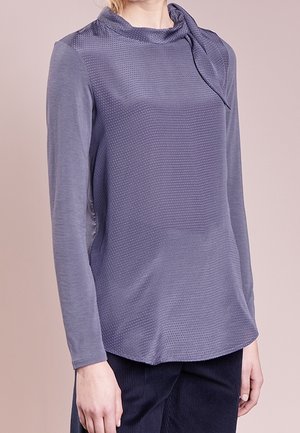 Femme portant un haut violet à manches longues avec une texture et un détail drapé au col, associé à un pantalon foncé sur un fond uni.