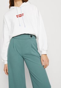 Kobieta nosząca białą bluzę Levi's z niebieskimi sznurkami i wysokimi, szerokimi spodniami w kolorze teal z czarnym guzikiem w talii.