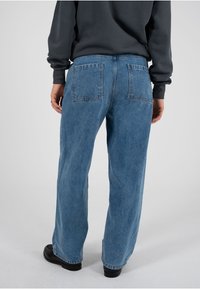 Jeans mit weitem Bein aus hellblauem Denim, mit Gesäßtaschen. Kombiniert mit einem dunklen Sweatshirt und schwarzen Schuhen. Glatte Textur und lockere Passform.