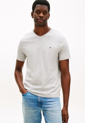 Hombre con camiseta de cuello en V gris claro con pequeño logo en el pecho y jeans azul claro, de pie con una mano en el bolsillo.