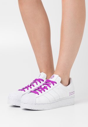 Białe sneakersy na platformie z fioletowymi sznurówkami, noszone na gołych nogach, z nakrapianą podeszwą i małym fioletowym napisem po zewnętrznej stronie.