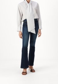 Randig långärmad skjorta med överdimensionerad fluga, mörka utsvängda jeans och bruna klacksandaler med nitar. Snygg silhuett, avslappnad textur.