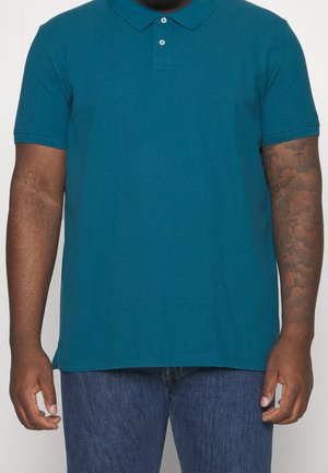 Mężczyzna w turkusowej koszulce polo z krótkim rękawem i niebieskich dżinsach, z tatuażem na prawym przedramieniu, stojący na tle białym.