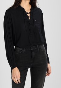 Black, long-sleeve blouse with a V-neck and lace-up front. Slight sheen on soft fabric, paired with dark jeans. Pocket on left chest.