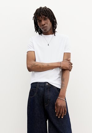 Tricou alb cu guler rotund, mâneci scurte, asortat cu blugi cu picioare largi de culoare albastru închis. Tatuaje notabile pe braț și brățară argintie pe încheietura mâinii.