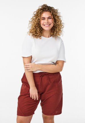 Lachende vrouw met krullend haar die een wit T-shirt met korte mouwen en roestkleurige shorts met trekkoord draagt, staand tegen een effen achtergrond.