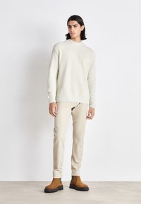 Pull en tricot beige clair avec ourlet côtelé, associé à un pantalon couleur crème et des bottines en suède marron avec des accents verts.