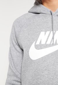 Sudadera gris con una tela suave y texturizada. Presenta un gran logo blanco de Nike en el frente y una capucha con cordón.