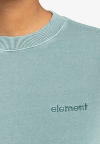 Sweat-shirt bleu clair avec un col rond côtelé et un logo brodé "element". Texture de tissu doux, vue rapprochée du col et de la zone de la poitrine.