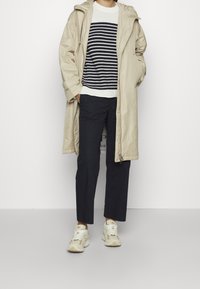 Beige lång kappa, vit och marinrandig stickad tröja, marinbyxor och ljusa sneakers. Kappan har en huva och dragkedja.