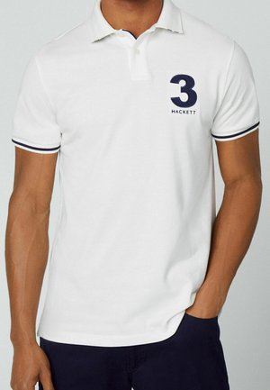 Mand iført hvid polo skjorte med marineblå kant på ærmerne og stort navyblåt "3 Hackett" logo på brystet, hænder i lommerne.