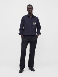 Homme debout les mains dans les poches, portant une chemise noire boutonnée avec un écusson sur la poitrine, pantalon noir et chaussures noires sur un fond clair uni.