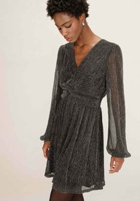 Donna che indossa un vestito lungo fino al ginocchio, nera e brillante, con un dettaglio a nodo attorcigliato in vita, in piedi contro uno sfondo chiaro e semplice.