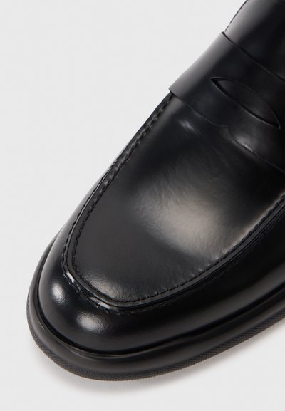 Svart läderloafer med en glansig finish, med en sydd tå och en rem över vaden. Slät textur och rundad form.