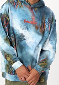 Persona che indossa un colorato hoodie Carlo Colucci con motivi astratti blu, arancione e giallo, mani unite in vita.