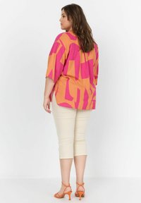 Korte mouwen, oversized shirt met een geometrisch patroon in roze en oranje, gecombineerd met crèmekleurige capri-broeken en oranje hakschoenen.