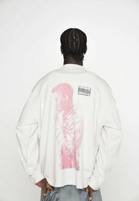 Biały długi rękaw sweatshirt z różową grafiką postaci na plecach oraz prostokątną czarną metką z białym tekstem.