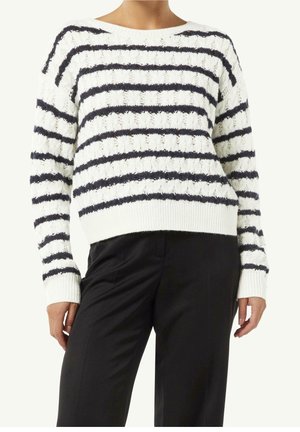 Femme portant un pull en maille blanc avec des rayures horizontales noires et un pantalon noir, montrée du cou jusqu'aux hanches.