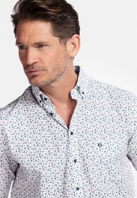 Chemise blanche à boutons avec un motif floral bleu et marron. Dotée d'un col à boutons et d'un logo sur la poitrine. Texture de tissu doux.