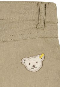 Tessuto beige con finitura testurizzata, dotato di una toppa con testa d'orso ricamata con un accento giallo, posizionata vicino all'orlo della vita.