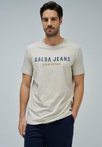 Camiseta de algodón beige con texto "SALSA JEANS" en azul y marrón, cuello redondo, mangas cortas y detalle de etiqueta lateral.