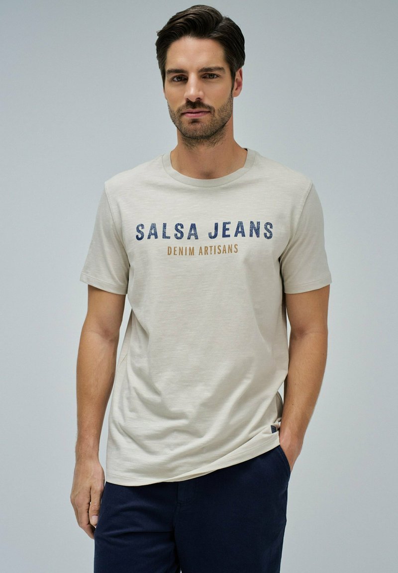 Camiseta de algodón beige con texto "SALSA JEANS" en azul y marrón, cuello redondo, mangas cortas y detalle de etiqueta lateral.