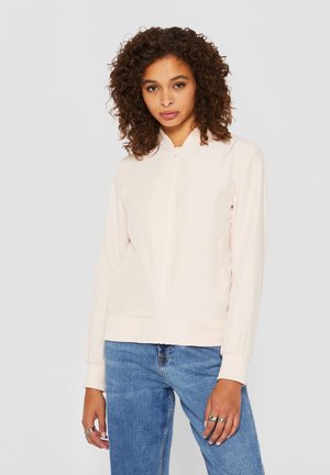 Vrouw met krullend bruin haar die een lichtbeige jas met rits en blauwe spijkerbroek draagt, staand tegen een effen grijze achtergrond.