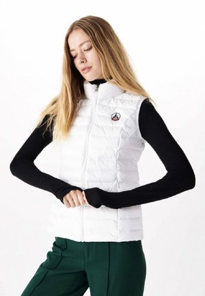 White quilted vest with high collar and front zipper, worn over a black long-sleeve top, featuring a small circular logo on the chest.
