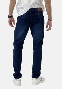 Jeans de mezclilla azul oscuro con corte ajustado, que cuentan con dos bolsillos traseros y un parche de cuero en la cintura. Llevados con zapatillas blancas.