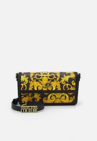 Czarna torebka na ramię z złotym kwiatowym wzorem, teksturowany materiał, prostokątny kształt, magnetyczne zapięcie i regulowany pasek z napisem "VERSACE JEANS COUTURE."