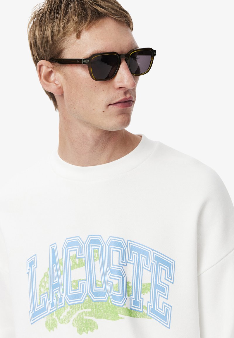 Felpa bianca con testo "LACOSTE" in blu e verde. Il modello indossa occhiali da sole scuri con una montatura in tartaruga testurizzata e lenti scure.
