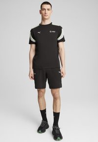 Musta urheilupaita, jossa on vihreitä ja valkoisia yksityiskohtia, yhdistetty mustiin shortsihin, joissa on tekstuuripinta ja taskut. Mustat kengät vihreillä/pohjilla.