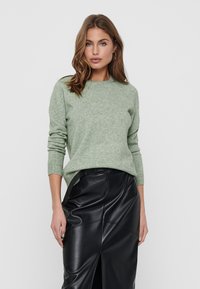 Pull en tricot vert clair à manches longues, col rond et poignets côtelés, associé à une jupe en cuir noire avec une fente sur le côté.