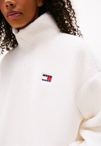 Witte fleecejas met een hoge kraag, voorzien van een klein, kleurrijk Tommy Jeans-logo aan de linkerkant. Zachte textuur en een relaxte pasvorm.