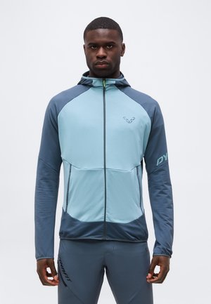 Atlétikus férfi, aki kék kapucnis, teljes cipzáras pulóvert és hozzá illő nadrágot visel, és egy sima fehér háttér előtt áll.