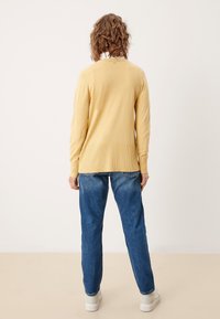 Osoba z kręconymi włosami, ubrana w jasną żółtą bluzę, niebieskie dżinsy i białe trampki, stojąca na tle jednolitego jasnego tła.