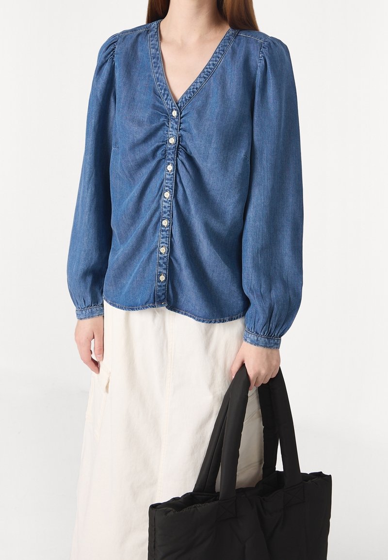 Femme portant un chemisier en denim bleu boutonné avec un devant froncé, jupe blanche, tenant un sac cabas noir matelassé par les anses.