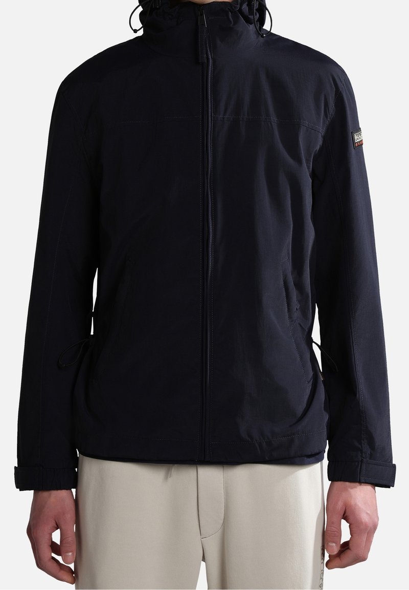 Chaqueta de color azul marino con cremallera frontal, capucha ajustable, puños elásticos y bolsillos laterales. Fabricada con un material ligero y resistente al agua.