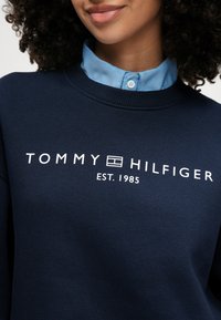 Ναυτικό βαμβακερό φούτερ με ριγέ λαιμόκοψη, featuring λογότυπο "TOMMY HILFIGER" σε λευκό χρώμα και κείμενο "EST. 1985". Κολάρο σε ανοιχτό μπλε χρώμα από κάτω.
