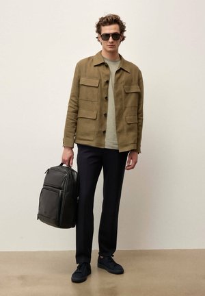 Vīrietis ar saulesbrillēm, olīvu krāsas jaku, gaiši pelēku kreklu, melnas bikses, melnas sporta kurpes, tur melnu ādas mugursomu pie sāniem.