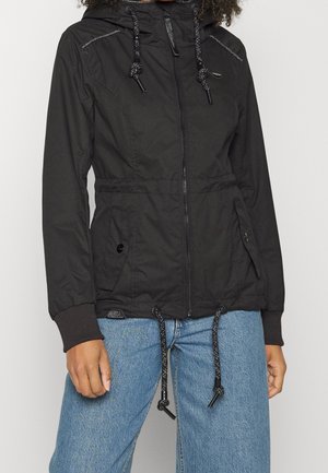 Kobieta nosząca czarną kurtkę z kapturem na zamek błyskawiczny z sznurkami, kieszeniami bocznymi na guziki i ściągaczami, w połączeniu z niebieskimi dżinsami.