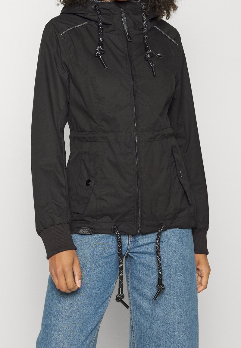 Femme portant une veste noire à capuche zippée avec cordons, poches latérales à boutons et poignets côtelés, associée à un jean bleu.